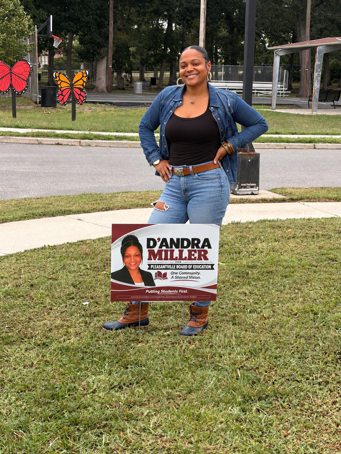 D’Andra Miller standing with her school board campaign sign at a neighborhood park